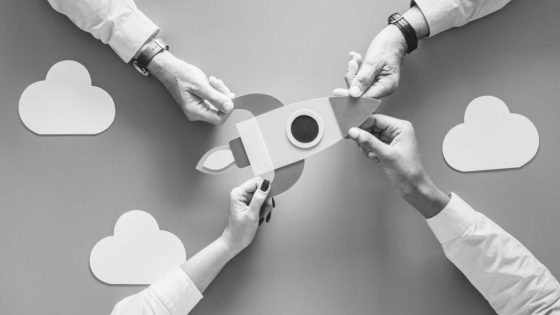 Four hands collaborating to assemble a paper rocket, symbolizing teamwork, innovation and entrepreneurship in launching a startup concept.